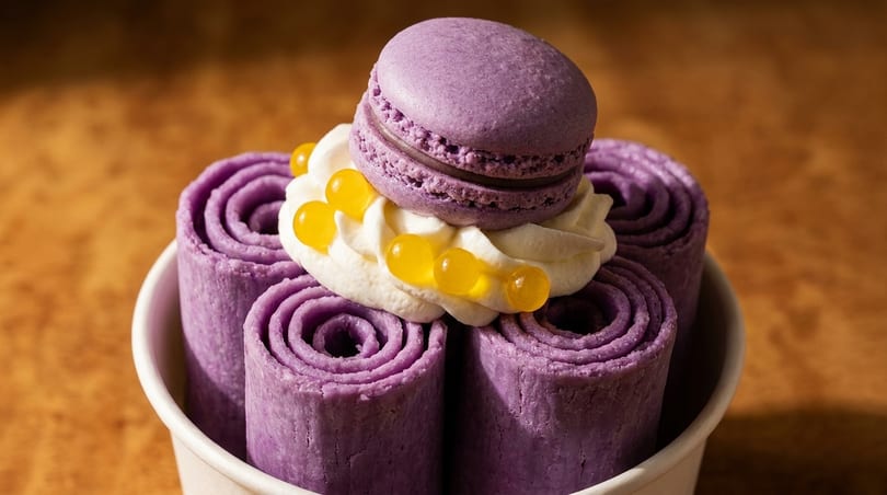 Purple rolled ice cream in a cup, topped with whipped cream, yellow boba, and a macaron.