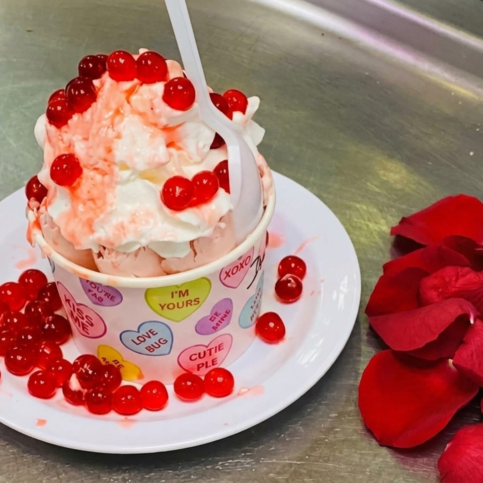 Pink ice cream with whipped cream and red boba in a heart-patterned cup, with rose petals.
