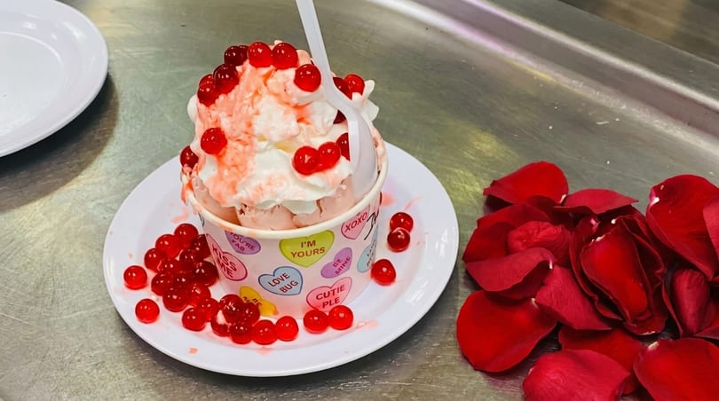 Pink ice cream with whipped cream and red boba in a heart-patterned cup, with rose petals.